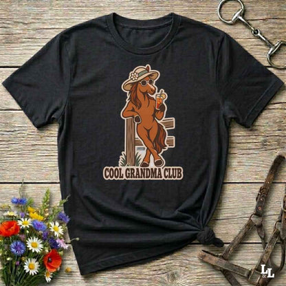 A black short-sleeve graphic T-shirt laid flat on a rustic wooden surface, featuring a cowboy horse illustration wearing a hat and sunglasses and holding a drink, with the text 'COOL GRANDMA CLUB' below. Decorative flowers and horse tack are placed around the shirt.