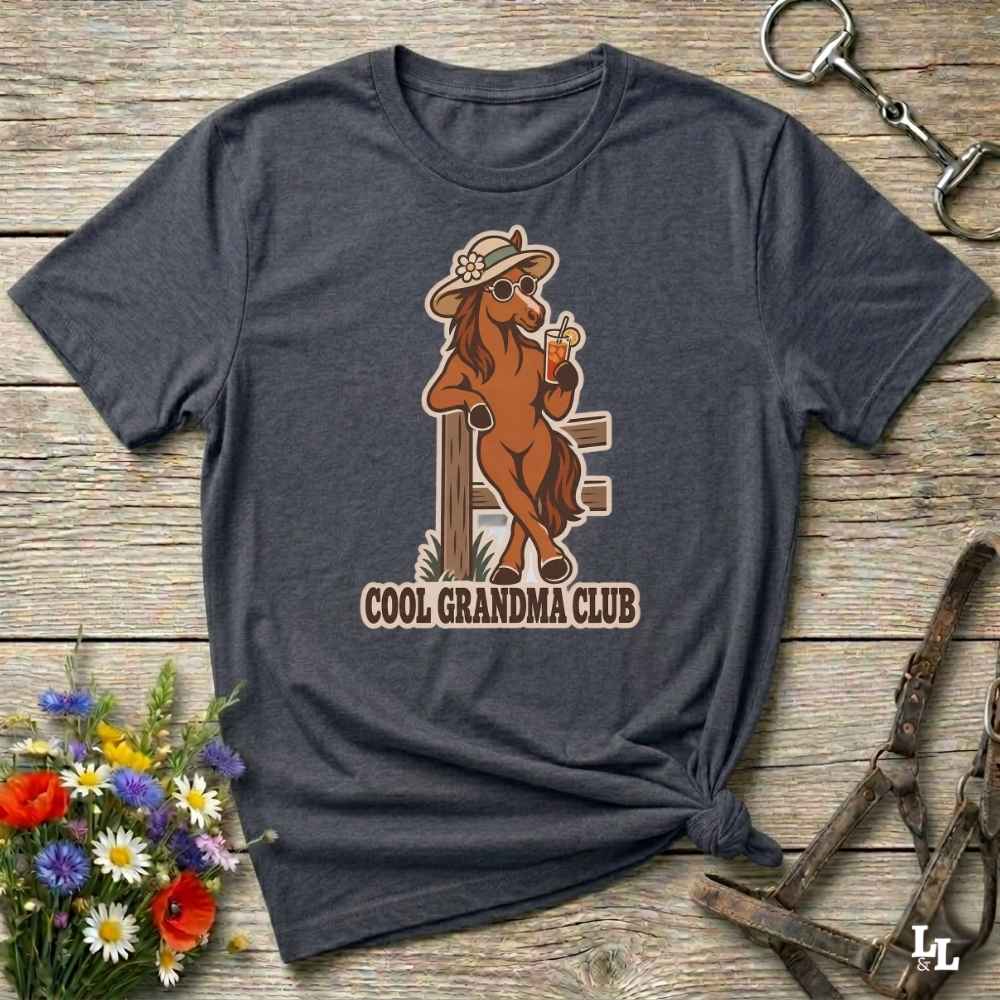A dark heather gray t-shirt laid flat on a rustic wooden surface, featuring a graphic of a cool brown horse wearing a hat and sunglasses and holding a drink, with the text 'COOL GRANDMA CLUB' underneath. The shirt is styled with equestrian accessories (a halter and a metal snap hook) and a small bouquet of wildflowers.