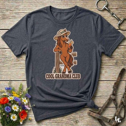 A dark heather gray t-shirt laid flat on a rustic wooden surface, featuring a graphic of a cool brown horse wearing a hat and sunglasses and holding a drink, with the text 'COOL GRANDMA CLUB' underneath. The shirt is styled with equestrian accessories (a halter and a metal snap hook) and a small bouquet of wildflowers.