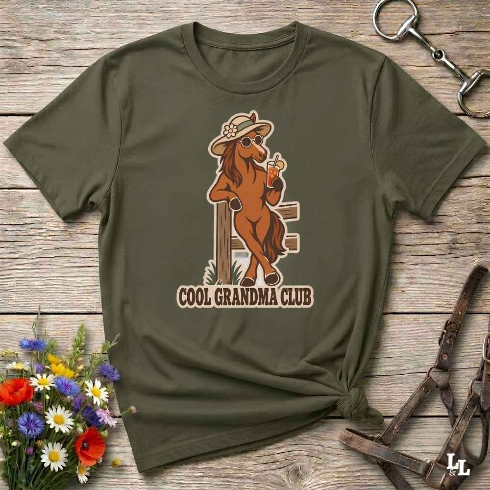 An olive green short-sleeve t-shirt with a graphic of a cowboy horse holding a beer and the text 'COOL GRANDMA CLUB', displayed on a rustic wooden background with flowers and horse tack.