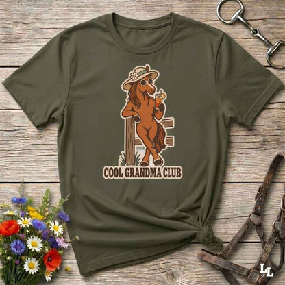 An olive green short-sleeve t-shirt with a graphic of a cowboy horse holding a beer and the text 'COOL GRANDMA CLUB', displayed on a rustic wooden background with flowers and horse tack.