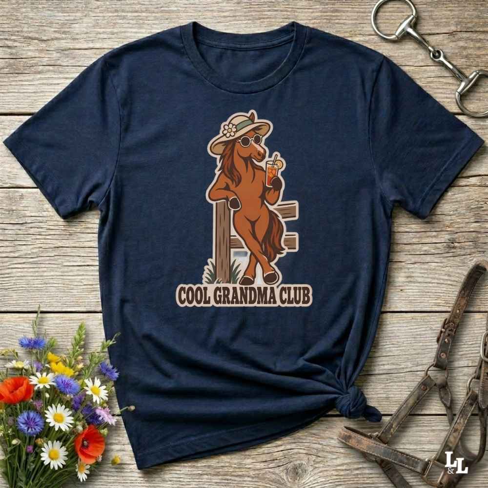 A navy blue graphic t-shirt featuring a retro illustration of a cool brown horse wearing a cowboy hat and sunglasses, holding a drink with the text 'COOL GRANDMA CLUB' underneath. The shirt is styled in a flat-lay on a rustic wooden background with equestrian accessories and a small bouquet of wildflowers.