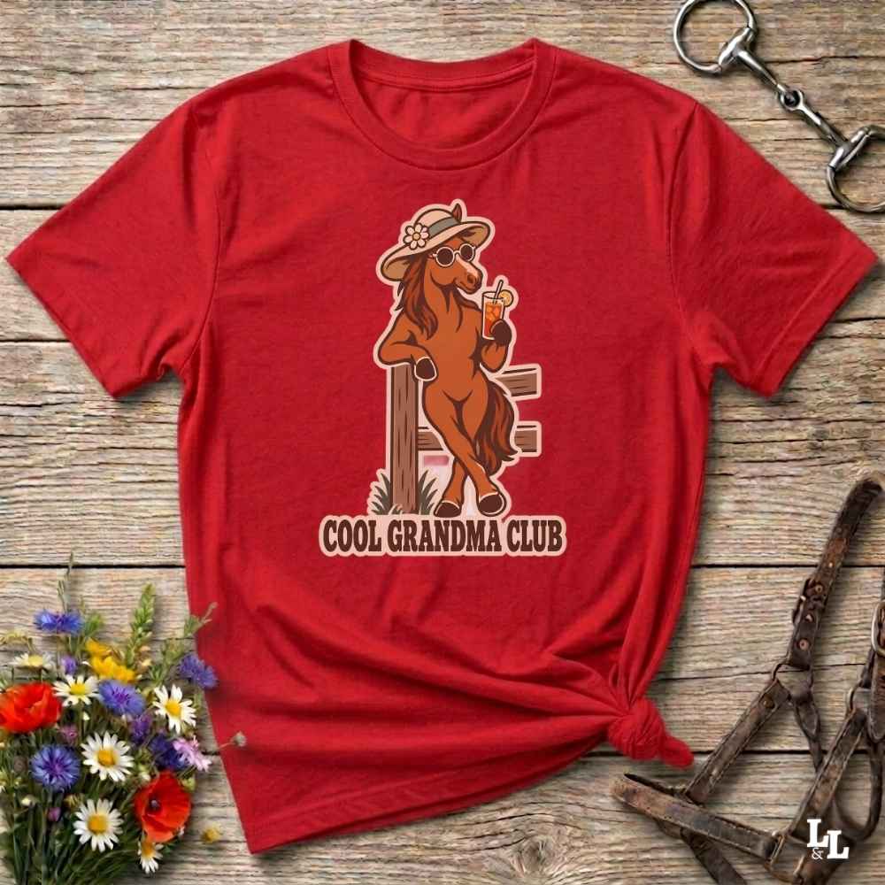 A red short-sleeve graphic T-shirt laid flat on a rustic wooden surface, styled with flowers, a horse bit, and a leather bridle. The shirt features a cartoon horse wearing a hat and sunglasses, holding a drink, with the text 'COOL GRANDMA CLUB' below.