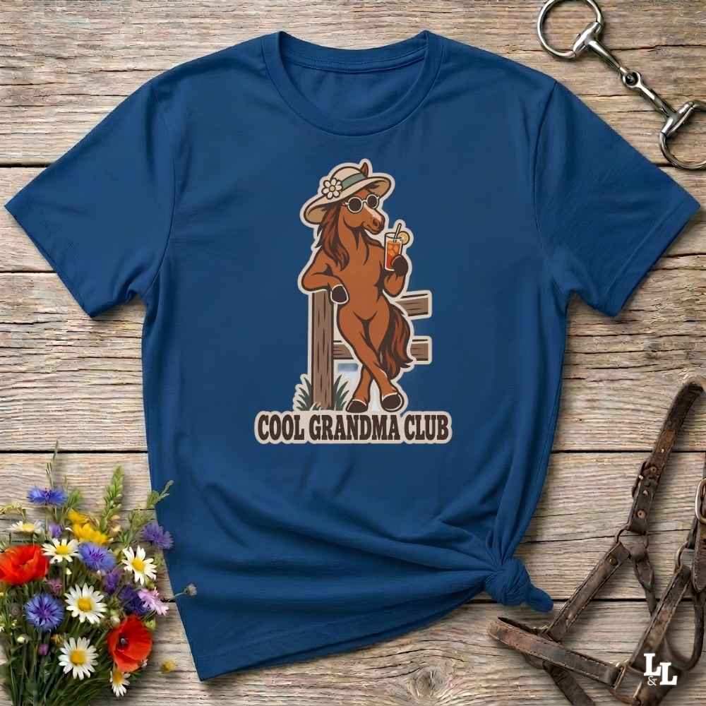 A navy blue short-sleeve t-shirt laid flat on a rustic wooden surface, featuring a cartoon graphic of a cool brown horse wearing a hat and sunglasses while holding a drink, with the text 'COOL GRANDMA CLUB' below. Decorative flowers and horse tack are placed around the shirt.