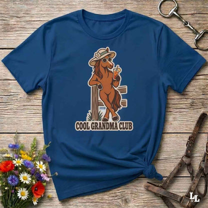 A navy blue short-sleeve t-shirt laid flat on a rustic wooden surface, featuring a cartoon graphic of a cool brown horse wearing a hat and sunglasses while holding a drink, with the text 'COOL GRANDMA CLUB' below. Decorative flowers and horse tack are placed around the shirt.