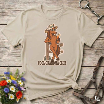 A beige short-sleeve t-shirt laid flat on a rustic wooden surface, featuring a graphic of a cool horse wearing a hat and sunglasses with the text 'COOL GRANDMA CLUB'. The shirt is styled with equestrian accessories and a small bouquet of wildflowers.