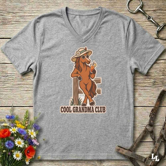 A heather gray V-neck t-shirt laid flat on a rustic wooden surface, featuring a cartoon graphic of a cool brown horse in a hat and sunglasses holding an orange drink, with the text 'COOL GRANDMA CLUB' below. Decorative flowers and equestrian tack are placed around the shirt.