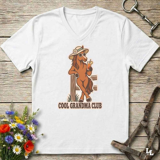 A white V-neck t-shirt laid flat on a rustic wooden surface, featuring a cartoon graphic of a cool brown horse wearing a hat and sunglasses while holding an orange drink. The text 'COOL GRANDMA CLUB' is printed below the graphic.