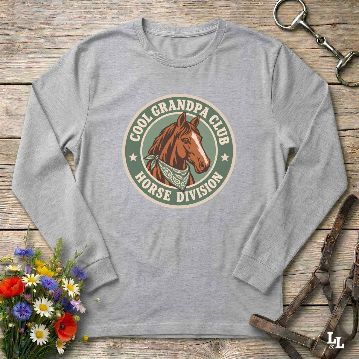 A long-sleeve crewneck t-shirt in grey, laid flat on a rustic wooden surface. The shirt features a circular graphic print in the center with a brown horse illustration and the text 'COOL GRANDPA CLUB HORSE DIVISION'.