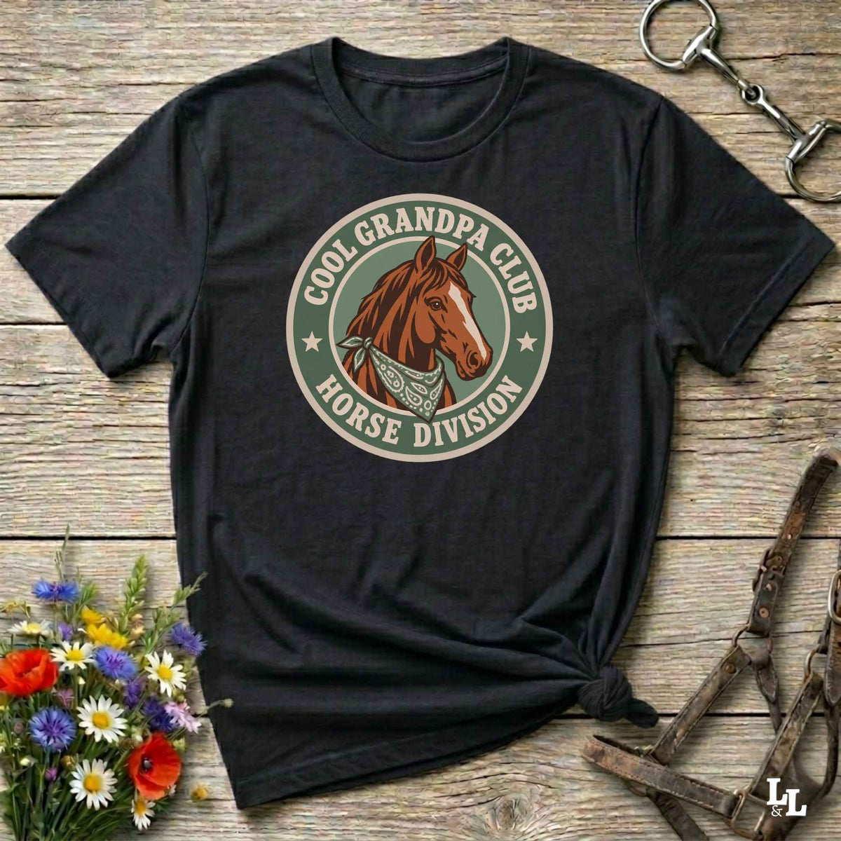 A black graphic t-shirt featuring a circular 'Cool Grandpa Club Horse Division' design with a brown horse illustration, styled on a rustic wooden background with equestrian accessories and flowers.