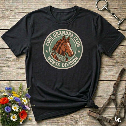 A black graphic t-shirt featuring a circular 'Cool Grandpa Club Horse Division' design with a brown horse illustration, styled on a rustic wooden background with equestrian accessories and flowers.