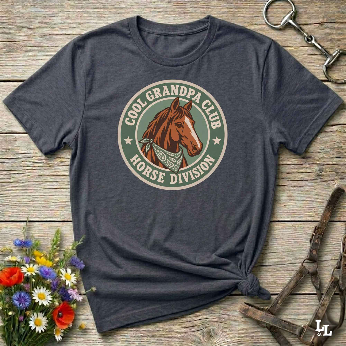 A dark heather gray graphic t-shirt laid flat on a rustic wooden surface, styled with equestrian accessories and a small bouquet of wildflowers. The shirt features a circular retro-style graphic with a brown horse illustration and the text 'COOL GRANDPA CLUB HORSE DIVISION'.