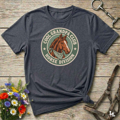 A dark heather gray graphic t-shirt laid flat on a rustic wooden surface, styled with equestrian accessories and a small bouquet of wildflowers. The shirt features a circular retro-style graphic with a brown horse illustration and the text 'COOL GRANDPA CLUB HORSE DIVISION'.
