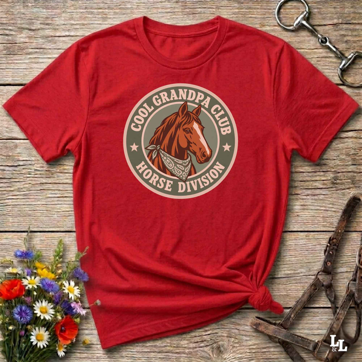 A red short-sleeve graphic T-shirt laid flat on a rustic wooden surface, styled with flowers, a horse bit, and a leather bridle. The shirt has a circular vintage-style chest graphic reading 'COOL GRANDPA CLUB HORSE DIVISION' around a brown horse illustration.