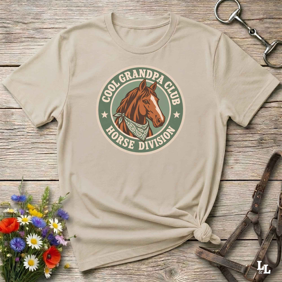 A beige graphic t-shirt with a 'Cool Grandpa Club Horse Division' logo featuring a brown horse, styled in a flat-lay on a rustic wooden background with equestrian accessories and flowers.