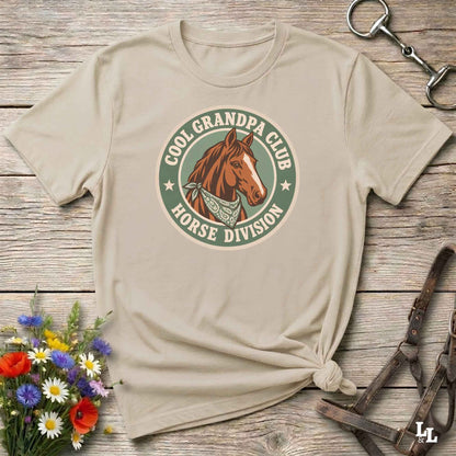 A beige graphic t-shirt with a 'Cool Grandpa Club Horse Division' logo featuring a brown horse, styled in a flat-lay on a rustic wooden background with equestrian accessories and flowers.