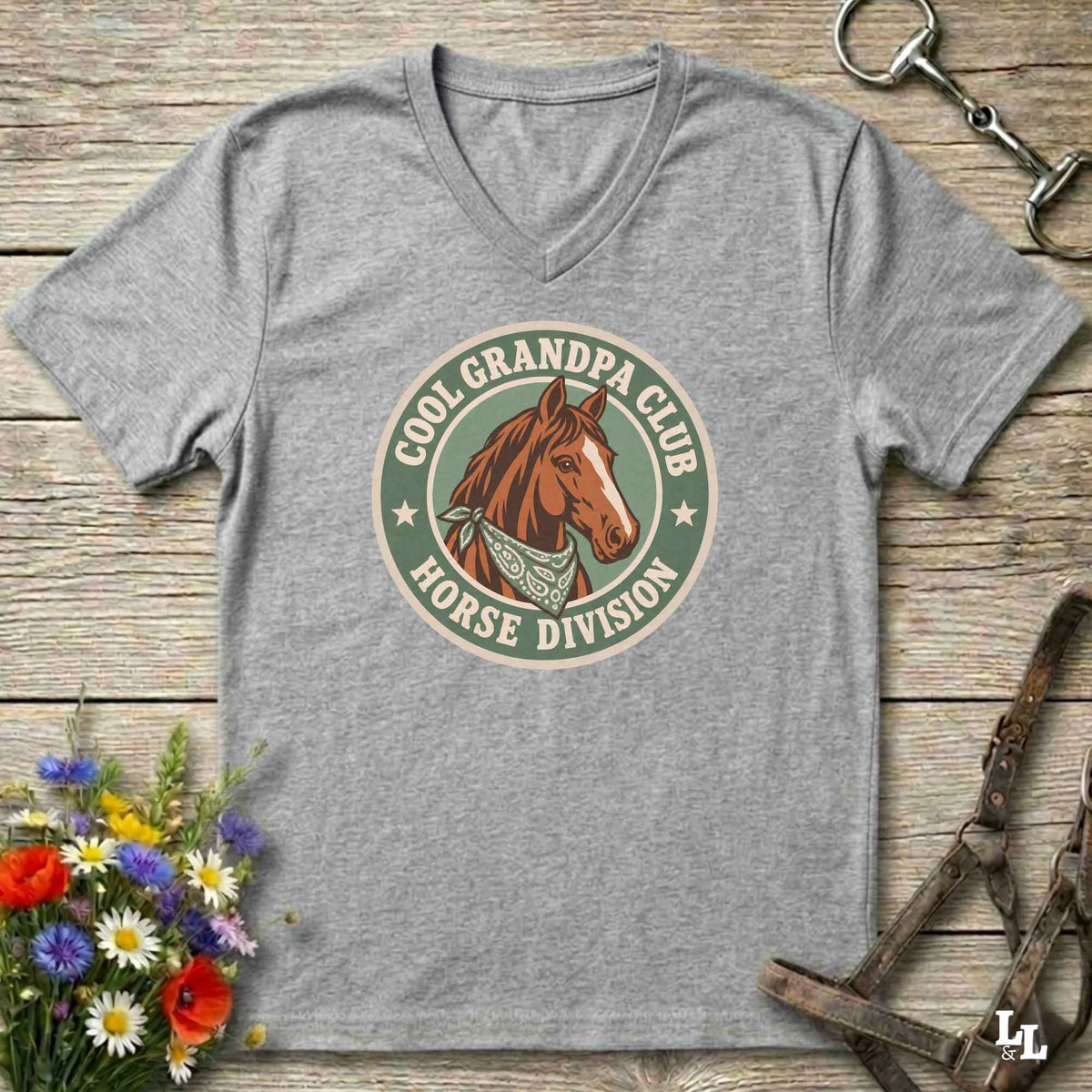 A heather gray v-neck t-shirt laid flat on a rustic wooden surface, featuring a retro-style circular graphic with a brown horse illustration and the text 'COOL GRANDPA CLUB HORSE DIVISION'. The shirt is styled with equestrian accessories (a halter and bit) and a small bouquet of colorful wildflowers.