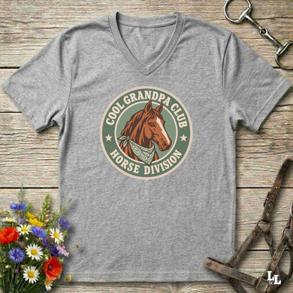 A heather gray v-neck t-shirt laid flat on a rustic wooden surface, featuring a retro-style circular graphic with a brown horse illustration and the text 'COOL GRANDPA CLUB HORSE DIVISION'. The shirt is styled with equestrian accessories (a halter and bit) and a small bouquet of colorful wildflowers.