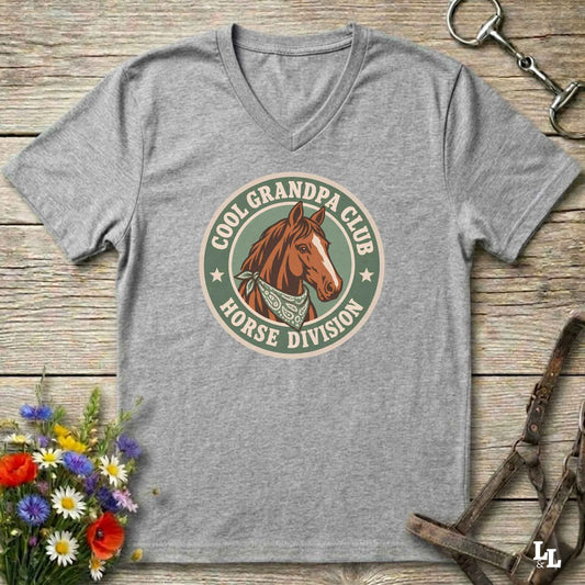 A heather gray v-neck t-shirt laid flat on a rustic wooden surface, featuring a retro-style circular graphic with a brown horse illustration and the text 'COOL GRANDPA CLUB HORSE DIVISION'. The shirt is styled with equestrian accessories (a halter and bit) and a small bouquet of colorful wildflowers.