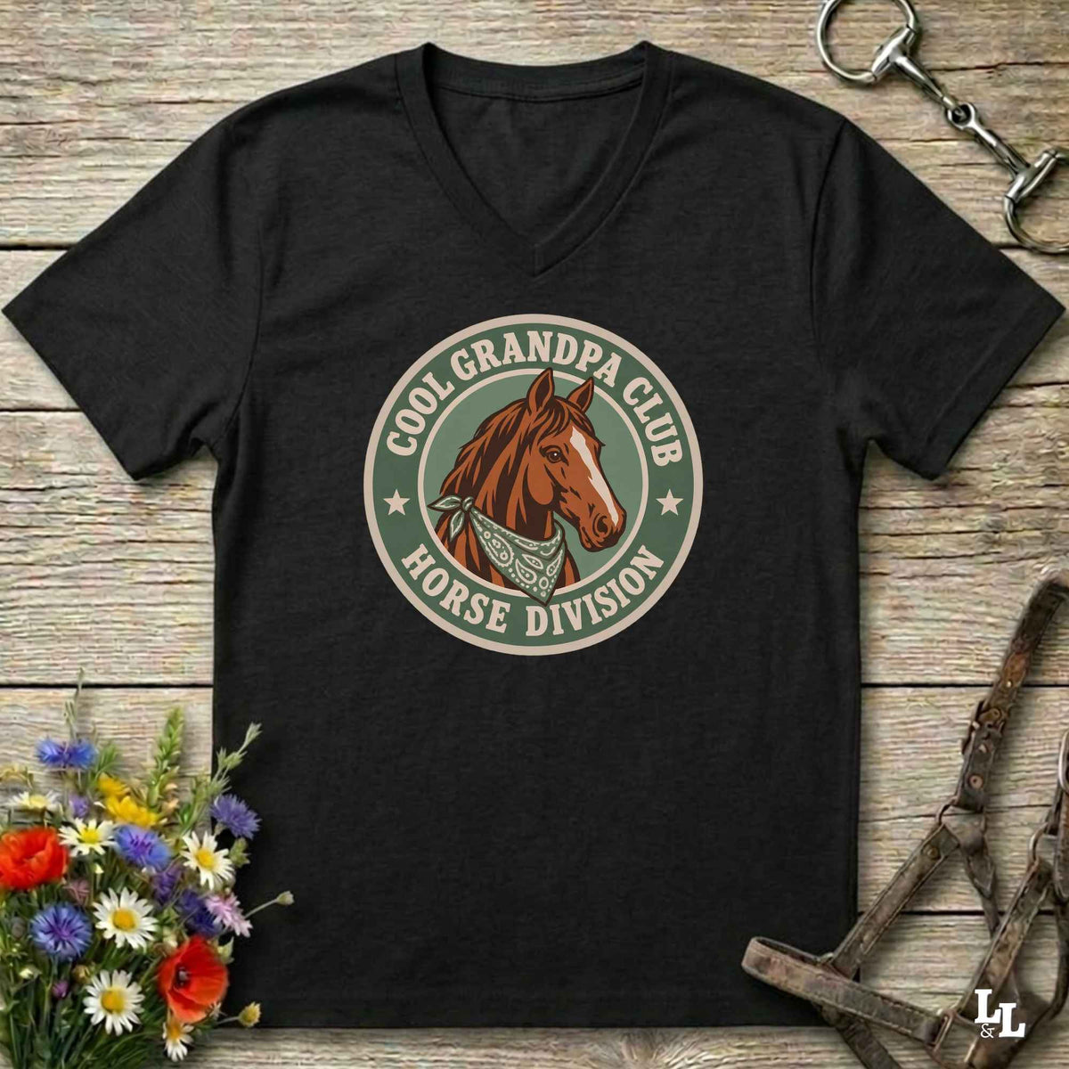 A black V-neck t-shirt laid flat on a rustic wooden surface, featuring a circular graphic of a brown horse head with the text 'COOL GRANDPA CLUB HORSE DIVISION'. The shirt is styled with equestrian props including a halter, tack, and a small bouquet of wildflowers.