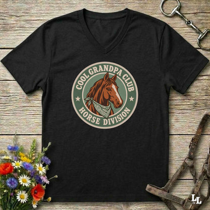 A black V-neck t-shirt laid flat on a rustic wooden surface, featuring a circular graphic of a brown horse head with the text 'COOL GRANDPA CLUB HORSE DIVISION'. The shirt is styled with equestrian props including a halter, tack, and a small bouquet of wildflowers.