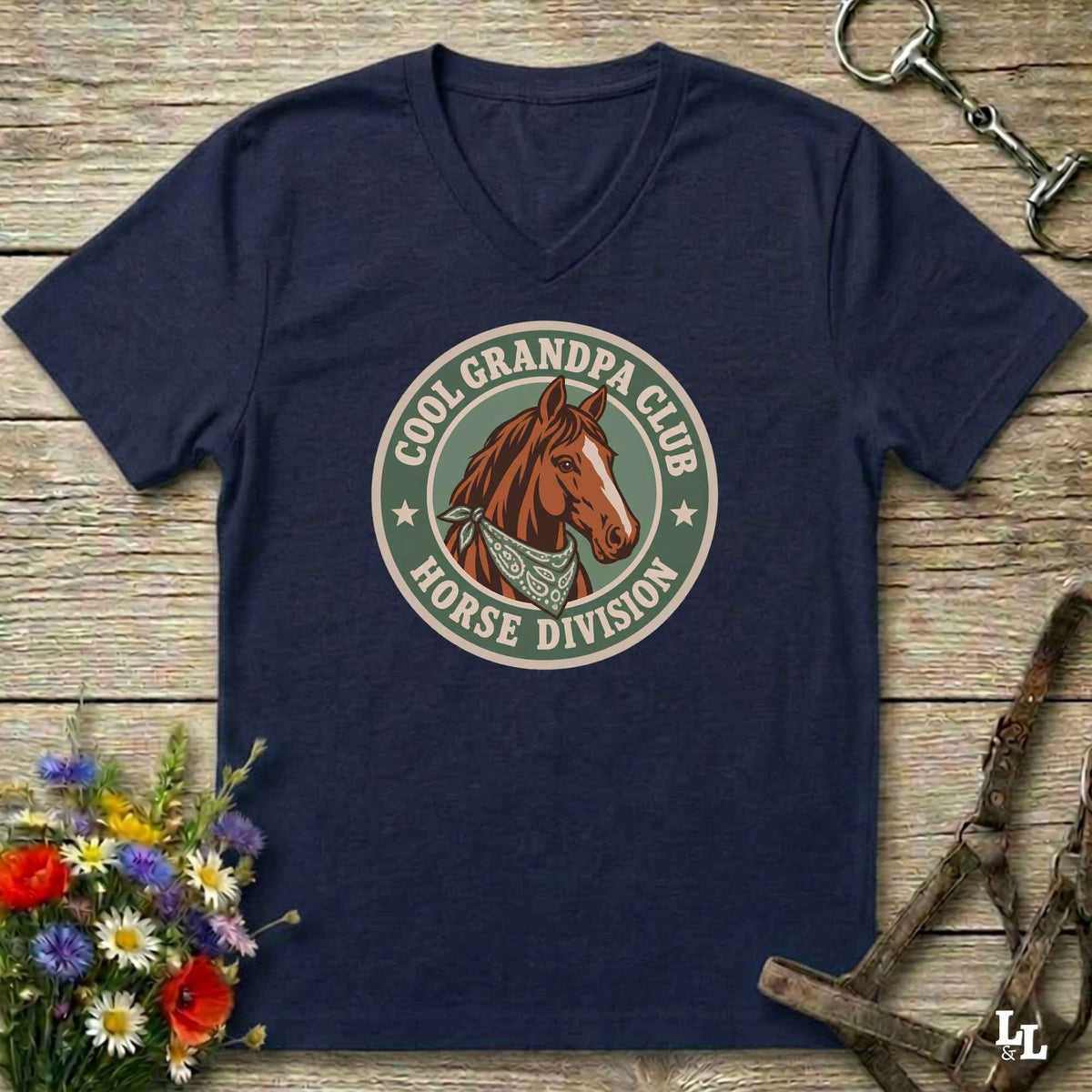 A navy blue v-neck t-shirt laid flat on a rustic wooden surface, featuring a circular graphic of a brown horse head with the text 'COOL GRANDPA CLUB HORSE DIVISION'. The shirt is styled with equestrian-themed props including a halter, tack, and a small bouquet of wildflowers.