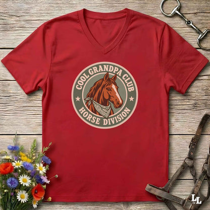 A red  v-neck t-shirt laid flat on a rustic wooden surface, featuring a circular graphic of a brown horse head with the text 'COOL GRANDPA CLUB HORSE DIVISION'. The shirt is styled with equestrian-themed props including a halter, tack, and a small bouquet of wildflowers.