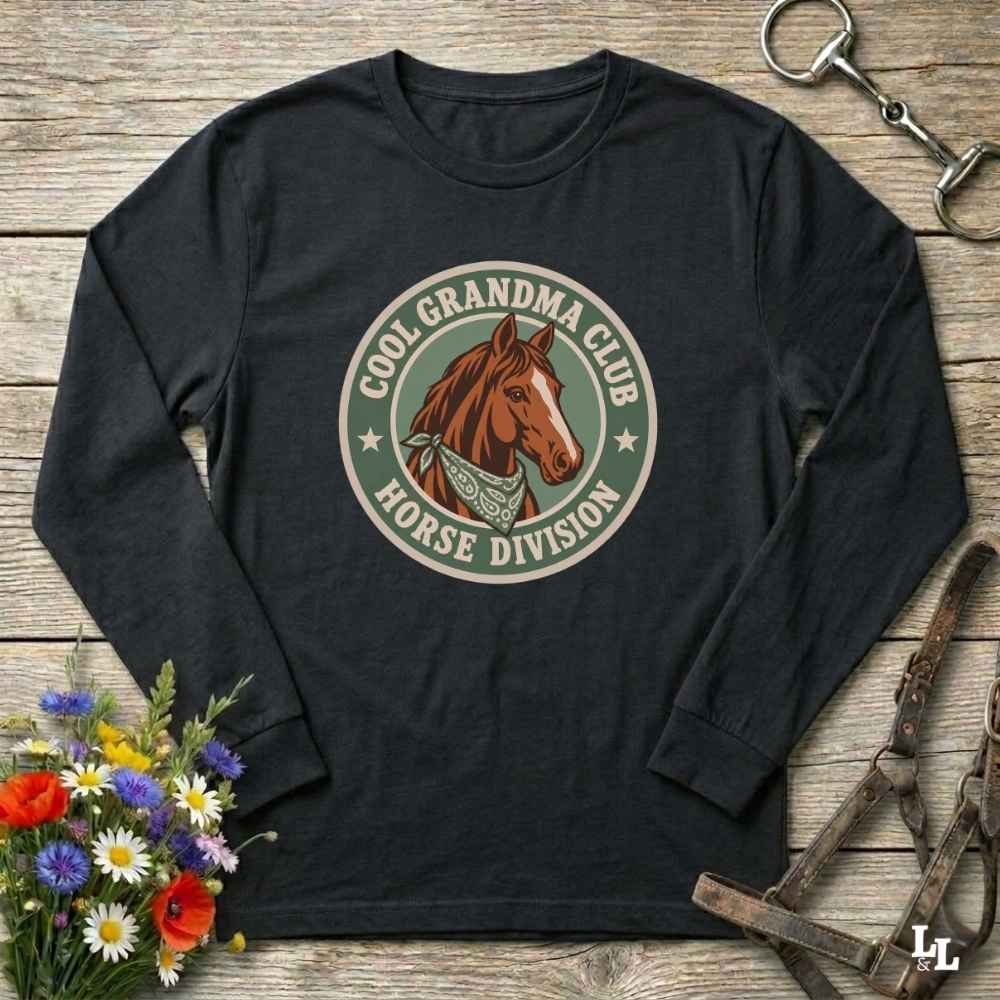 A black long-sleeve t-shirt with a circular graphic on the chest featuring a brown horse and the text 'COOL GRANDMA CLUB HORSE DIVISION'. The shirt is displayed in a flat-lay arrangement on a rustic wooden background, styled with a bouquet of colorful wildflowers, a horse bit, and a leather bridle.