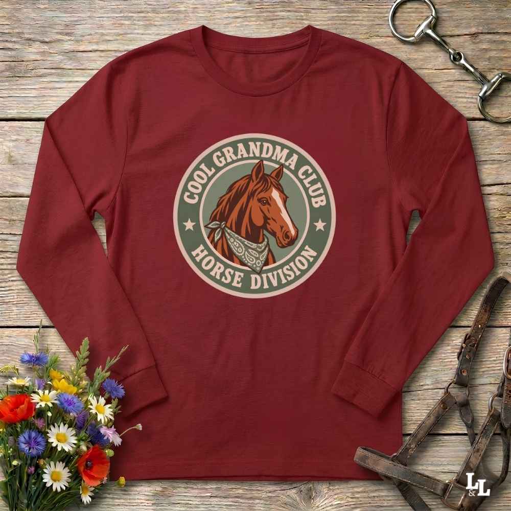 A cardinal red long-sleeve t-shirt with a circular graphic on the chest featuring a brown horse and the text 'COOL GRANDMA CLUB HORSE DIVISION'. The shirt is displayed in a flat-lay arrangement on a rustic wooden background, styled with a bouquet of colorful wildflowers, a horse bit, and a leather bridle.