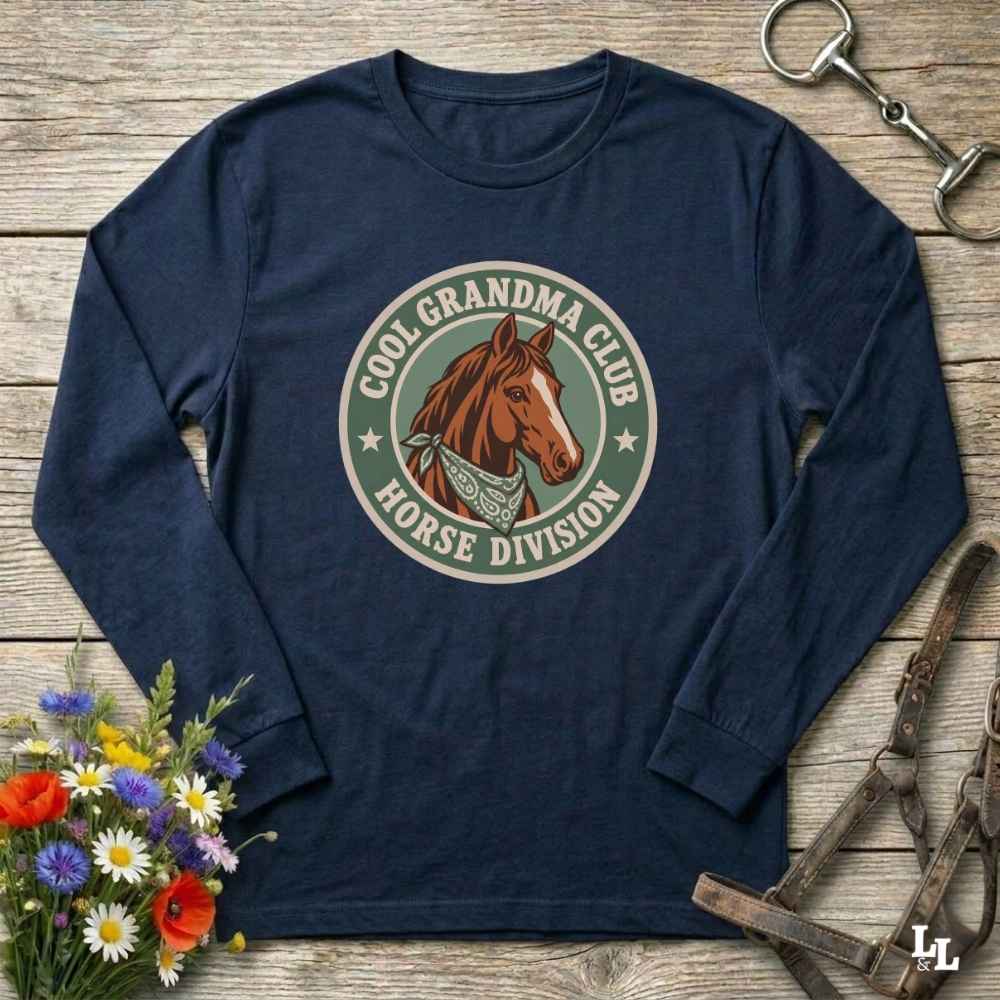 A blue  long-sleeve t-shirt with a circular graphic on the chest featuring a brown horse and the text 'COOL GRANDMA CLUB HORSE DIVISION'. The shirt is displayed in a flat-lay arrangement on a rustic wooden background, styled with a bouquet of colorful wildflowers, a horse bit, and a leather bridle.