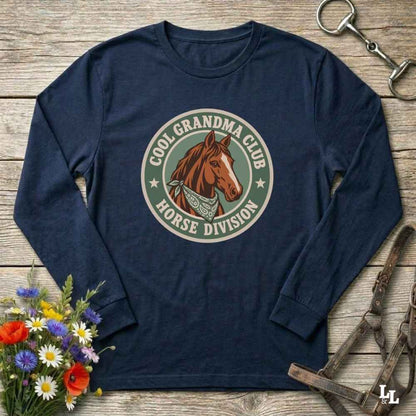 A blue  long-sleeve t-shirt with a circular graphic on the chest featuring a brown horse and the text 'COOL GRANDMA CLUB HORSE DIVISION'. The shirt is displayed in a flat-lay arrangement on a rustic wooden background, styled with a bouquet of colorful wildflowers, a horse bit, and a leather bridle.