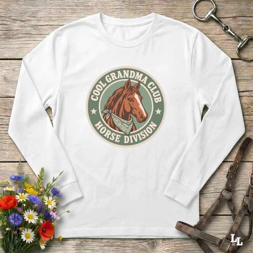 A white long-sleeve crewneck t-shirt with a circular graphic on the chest reading 'COOL GRANDMA CLUB HORSE DIVISION' around a brown horse illustration, styled flat-lay on a rustic wooden background with flowers and horse tack props.