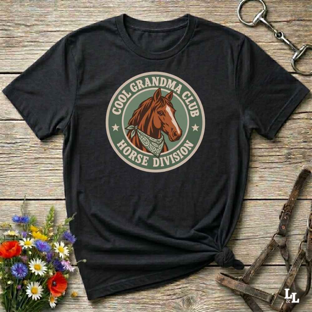 A black short-sleeve t-shirt with a circular graphic on the chest featuring a brown horse and the text 'COOL GRANDMA CLUB HORSE DIVISION'. The shirt is styled in a flat-lay on a rustic wooden background with equestrian accessories and flowers.
