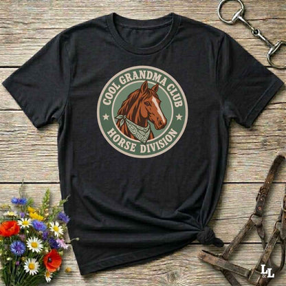 A black short-sleeve t-shirt with a circular graphic on the chest featuring a brown horse and the text 'COOL GRANDMA CLUB HORSE DIVISION'. The shirt is styled in a flat-lay on a rustic wooden background with equestrian accessories and flowers.