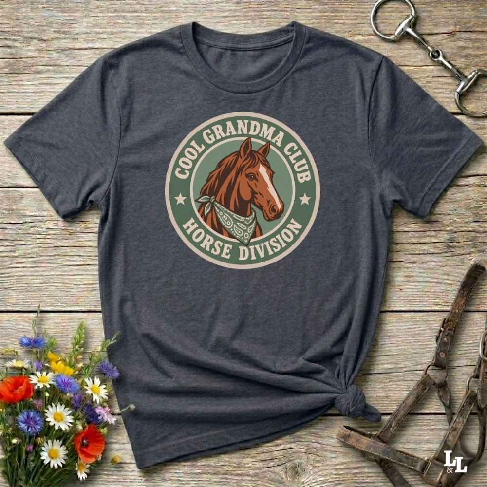 A dark heather gray graphic t-shirt laid flat on a rustic wooden surface, styled with equestrian accessories and a small bouquet of wildflowers. The shirt features a circular retro-style graphic with a brown horse illustration and the text 'COOL GRANDPA CLUB HORSE DIVISION'.