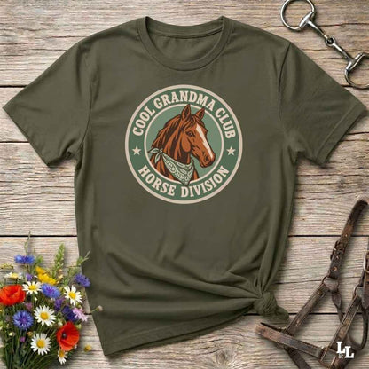 A green short-sleeve t-shirt with a circular graphic on the chest featuring a brown horse and the text 'COOL GRANDMA CLUB HORSE DIVISION'. The shirt is styled in a flat-lay on a rustic wooden background with equestrian accessories and flowers.