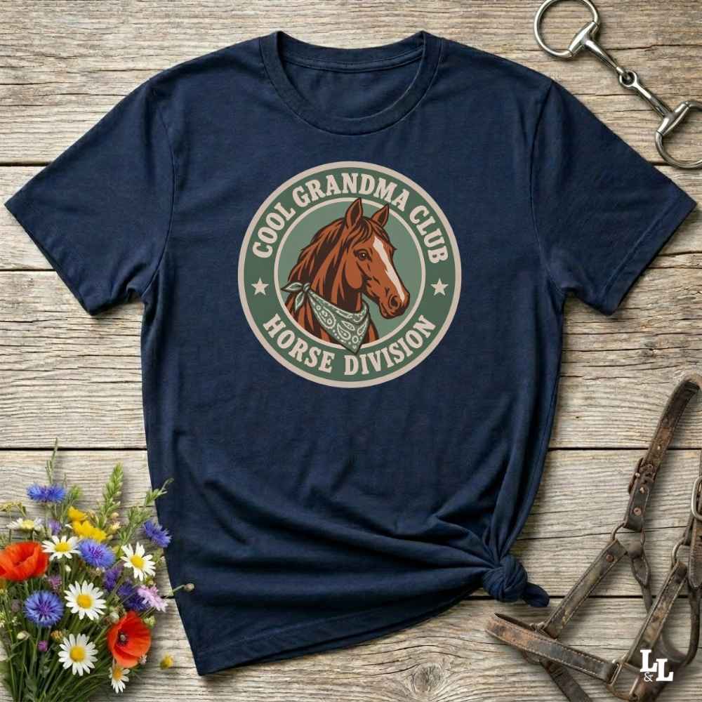 A navy blue short-sleeve t-shirt with a circular graphic on the chest featuring a brown horse and the text 'COOL GRANDMA CLUB HORSE DIVISION'. The shirt is styled in a flat-lay on a rustic wooden background with equestrian accessories and flowers.