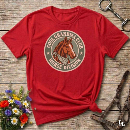 A red short-sleeve graphic T-shirt laid flat on a rustic wooden surface, styled with flowers, a horse bit, and a halter. The shirt features a circular vintage-style graphic reading 'COOL GRANDMA CLUB HORSE DIVISION' around an illustration of a brown horse.