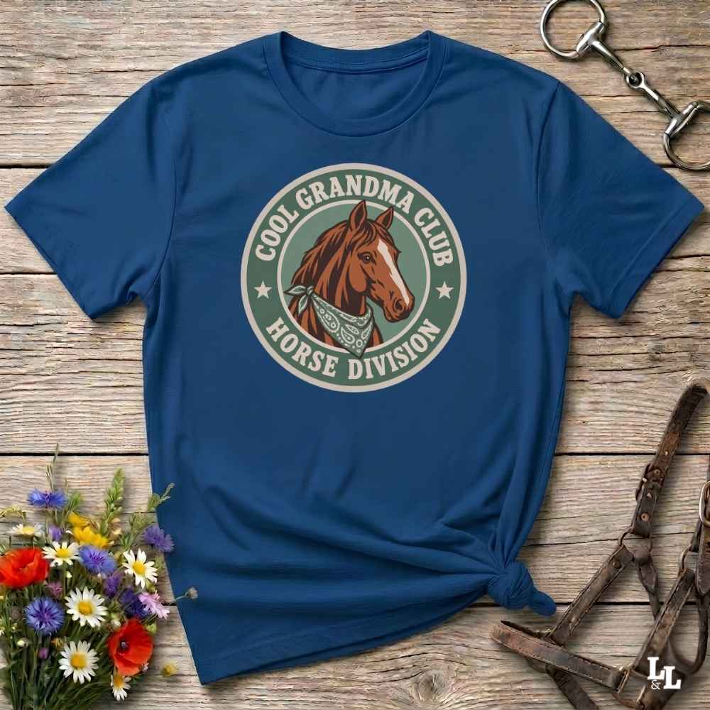A blue short-sleeve t-shirt with a circular graphic on the chest featuring a brown horse and the text 'COOL GRANDMA CLUB HORSE DIVISION'. The shirt is styled in a flat-lay on a rustic wooden background with equestrian accessories and flowers.