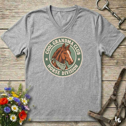 A heather gray v-neck t-shirt laid flat on a rustic wooden surface, featuring a retro-style circular graphic with a brown horse illustration and the text 'COOL GRANDPA CLUB HORSE DIVISION'. The shirt is styled with equestrian accessories (a halter and bit) and a small bouquet of colorful wildflowers.