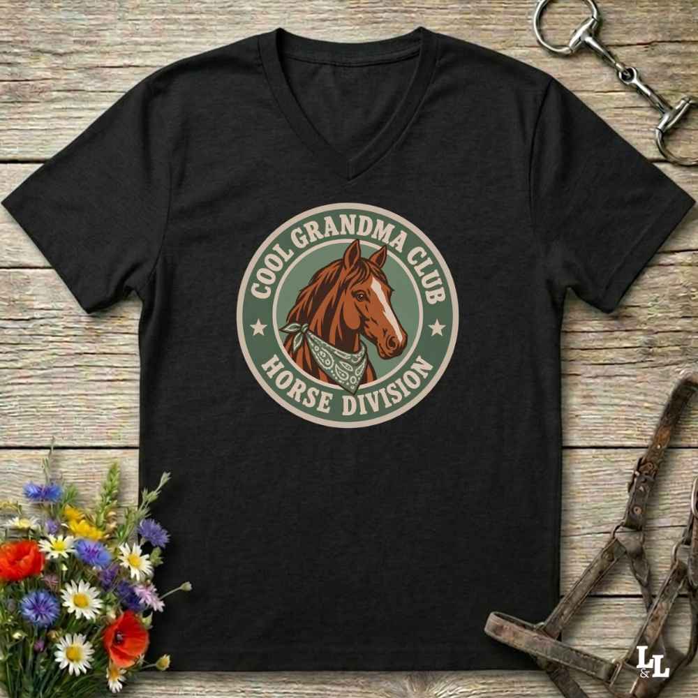 A black V-neck t-shirt with a retro-style circular graphic reading 'COOL GRANDMA CLUB HORSE DIVISION' around a brown horse illustration, laid flat on a rustic wood background with flowers and horse tack props.