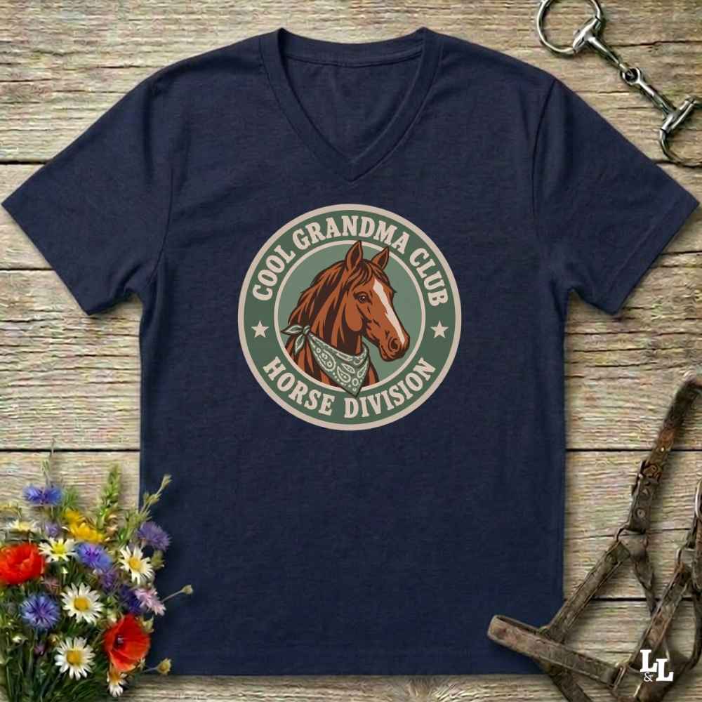 A navy blue v-neck t-shirt laid flat on a rustic wooden surface, featuring a circular graphic of a brown horse head with the text 'COOL GRANDPA CLUB HORSE DIVISION'. The shirt is styled with equestrian-themed props including a halter, tack, and a small bouquet of wildflowers.