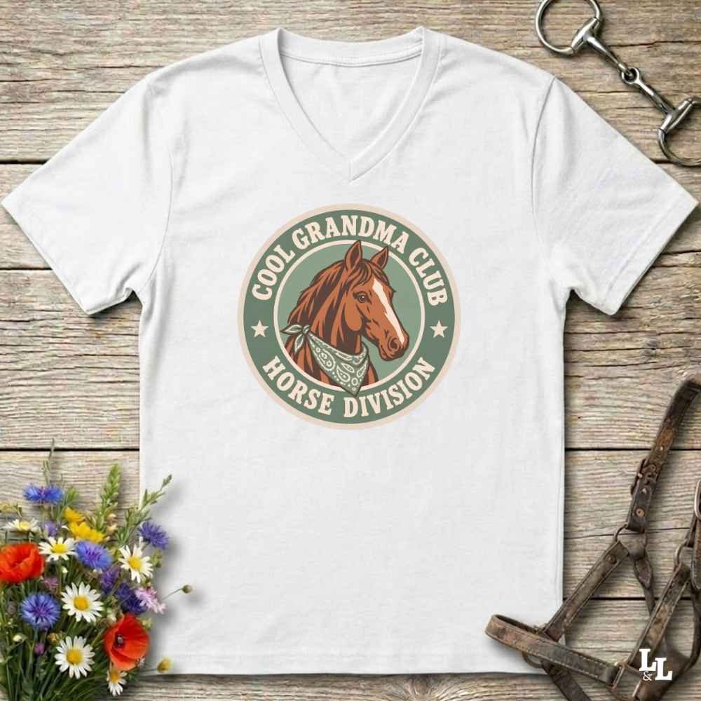 A white v-neck t-shirt laid flat on a rustic wooden surface, featuring a retro-style circular graphic with a brown horse illustration and the text 'COOL GRANDPA CLUB HORSE DIVISION'. The shirt is styled with equestrian accessories (a halter and bit) and a small bouquet of colorful wildflowers.