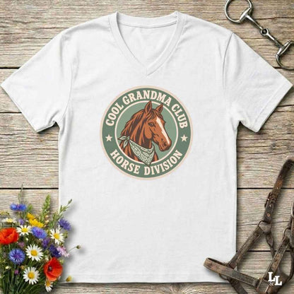 A white v-neck t-shirt laid flat on a rustic wooden surface, featuring a retro-style circular graphic with a brown horse illustration and the text 'COOL GRANDPA CLUB HORSE DIVISION'. The shirt is styled with equestrian accessories (a halter and bit) and a small bouquet of colorful wildflowers.