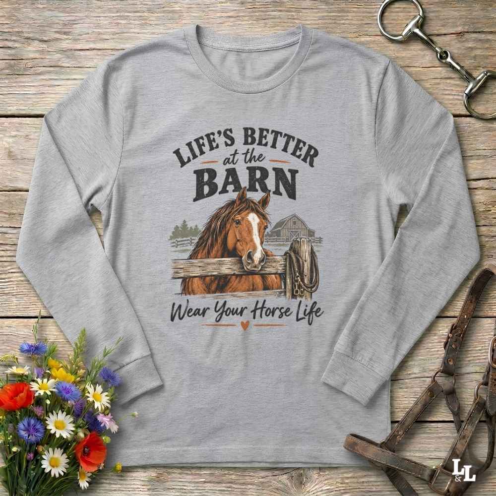 A light gray long-sleeve crewneck t-shirt with a graphic print of a brown horse and the text 'LIFE'S BETTER at the BARN Wear Your Horse Life', laid flat on a rustic wooden background with equestrian accessories and flowers.