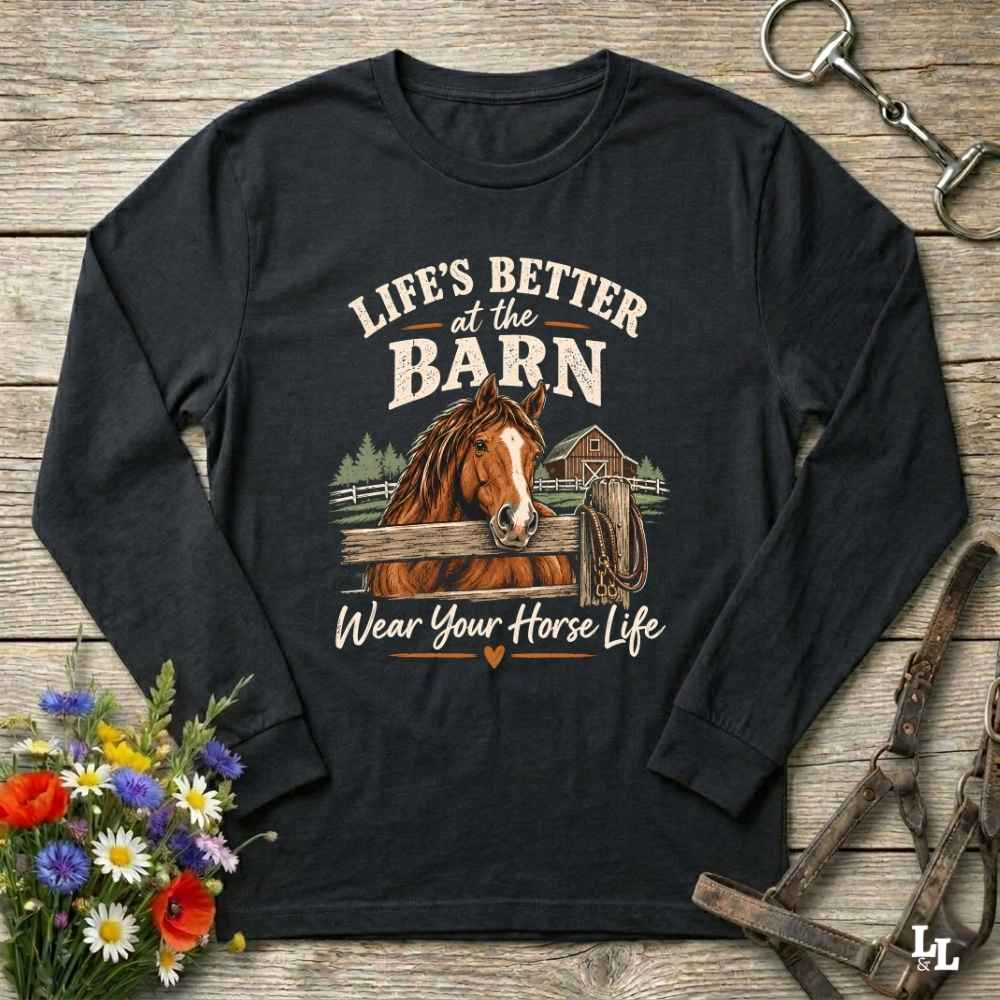A black long-sleeve t-shirt with a graphic print of a brown horse and the text 'LIFE'S BETTER at the BARN Wear Your Horse Life', displayed on a rustic wooden background with flowers and horse tack props.