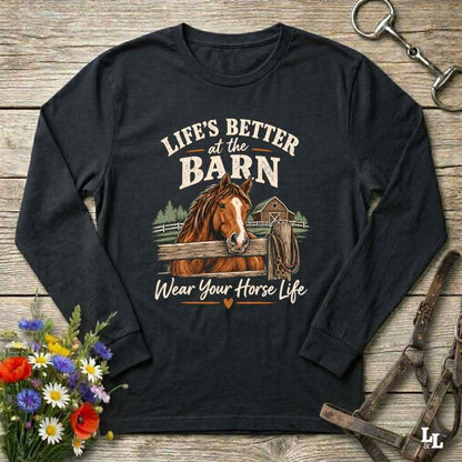 A black long-sleeve t-shirt with a graphic print of a brown horse and the text 'LIFE'S BETTER at the BARN Wear Your Horse Life', displayed on a rustic wooden background with flowers and horse tack props.