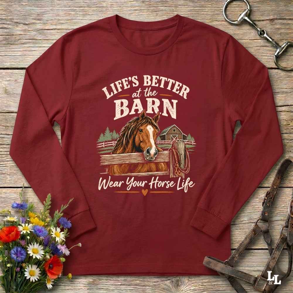 A long-sleeve crewneck t-shirt in maroon red, laid flat on a rustic wooden background. The shirt features a graphic print of a brown horse in a barn with the text 'LIFE'S BETTER at the BARN Wear Your Horse Life'.
