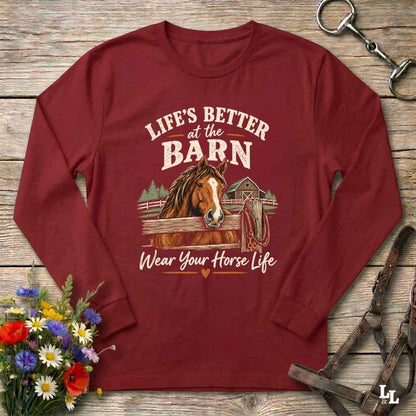 A long-sleeve crewneck t-shirt in maroon red, laid flat on a rustic wooden background. The shirt features a graphic print of a brown horse in a barn with the text 'LIFE'S BETTER at the BARN Wear Your Horse Life'.