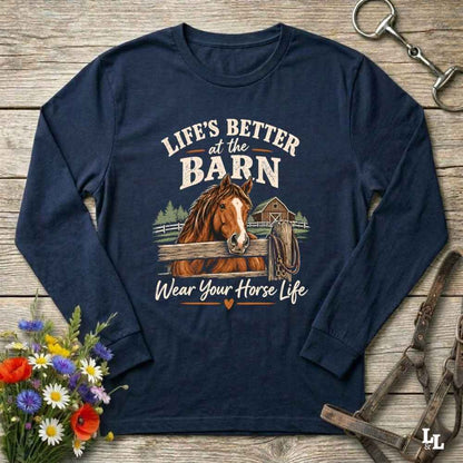 A navy blue long-sleeve crewneck t-shirt with a graphic print of a brown horse and the text 'LIFE'S BETTER at the BARN Wear Your Horse Life'. The shirt is displayed in a flat-lay arrangement on a rustic wooden background, styled with a bouquet of wildflowers, a horse bit, and a leather bridle.